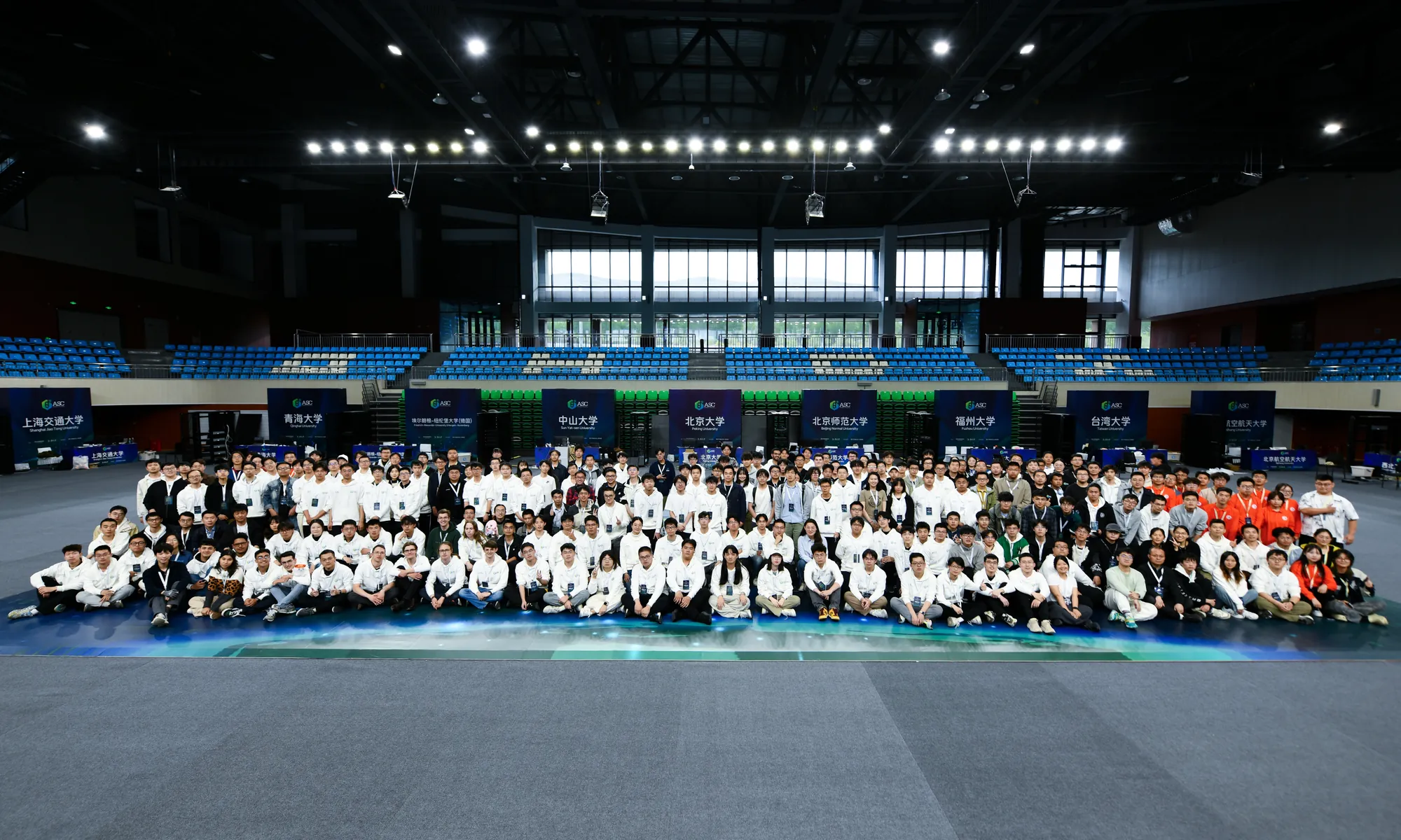 Several hundred competitors sitting on the floor of the finals venue for a group photo, with the universities' booths behind them