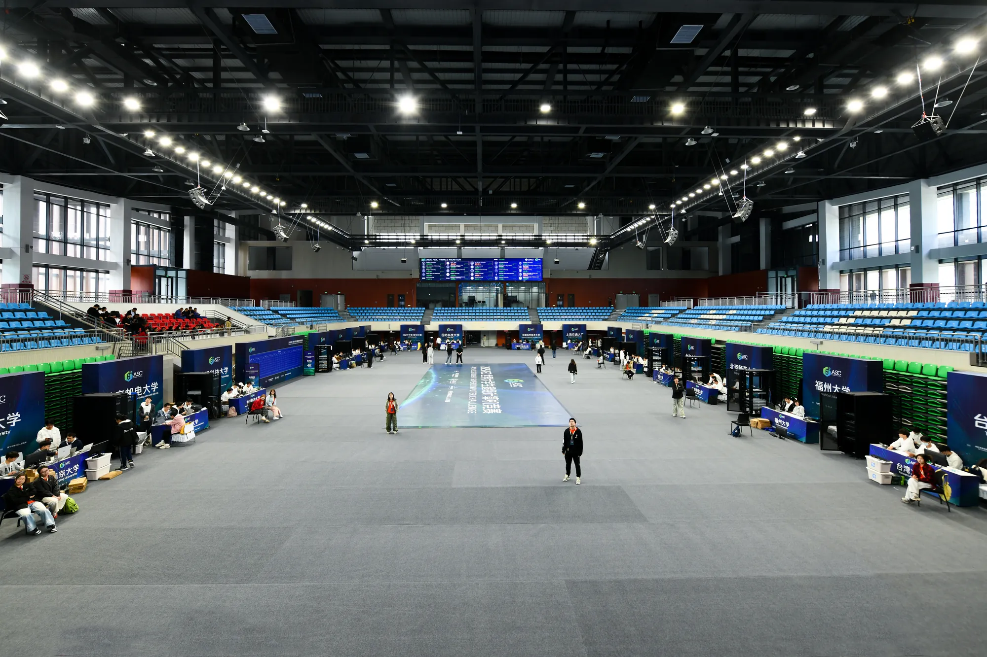 The finals venue seen from the stands, each university's racks and booths lining the edges, a live power-draw screen at the far end